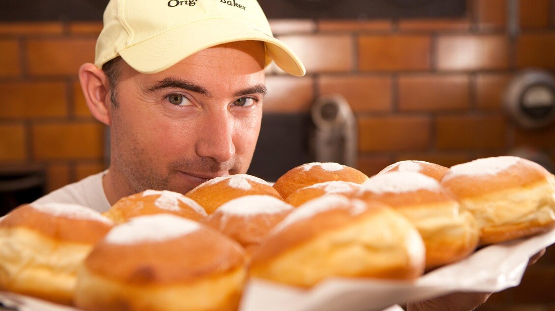 Ein Mann hält ein Tablett mit frischen Hefegebäck. Die Teilchen sind goldbraun und mit Puderzucker bestäubert. | © TV Region Graz - Harry Schiffer