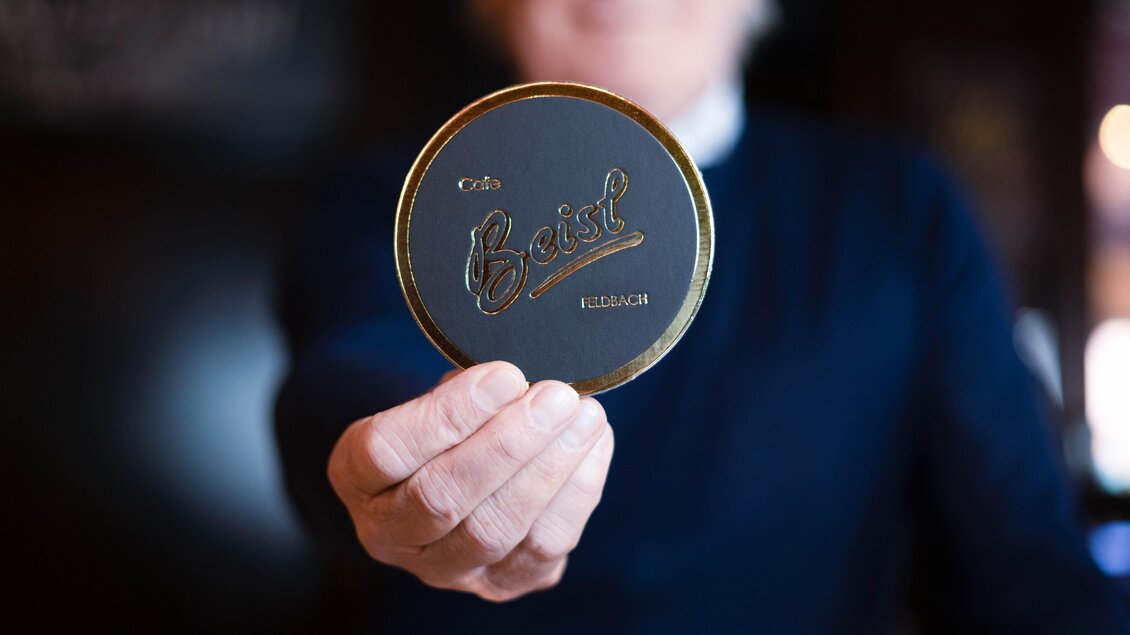 Eine Person hält einen runden Bierdeckel mit dem Schriftzug "Beisl" in der Hand. Der Hintergrund ist dunkel und unscharf. | © Cafe Beisl