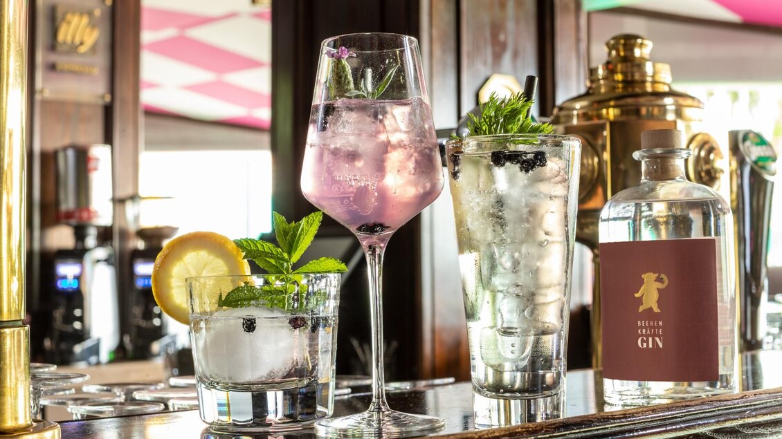 Drei Cocktails stehen auf der Bar, einer davon in einem eleganten Glas mit lila Getränk und frischen Minzblättern. Im Hintergrund ist eine stilvolle Bar mit bunten Akzenten zu sehen. | © Café Beisl Feldbach