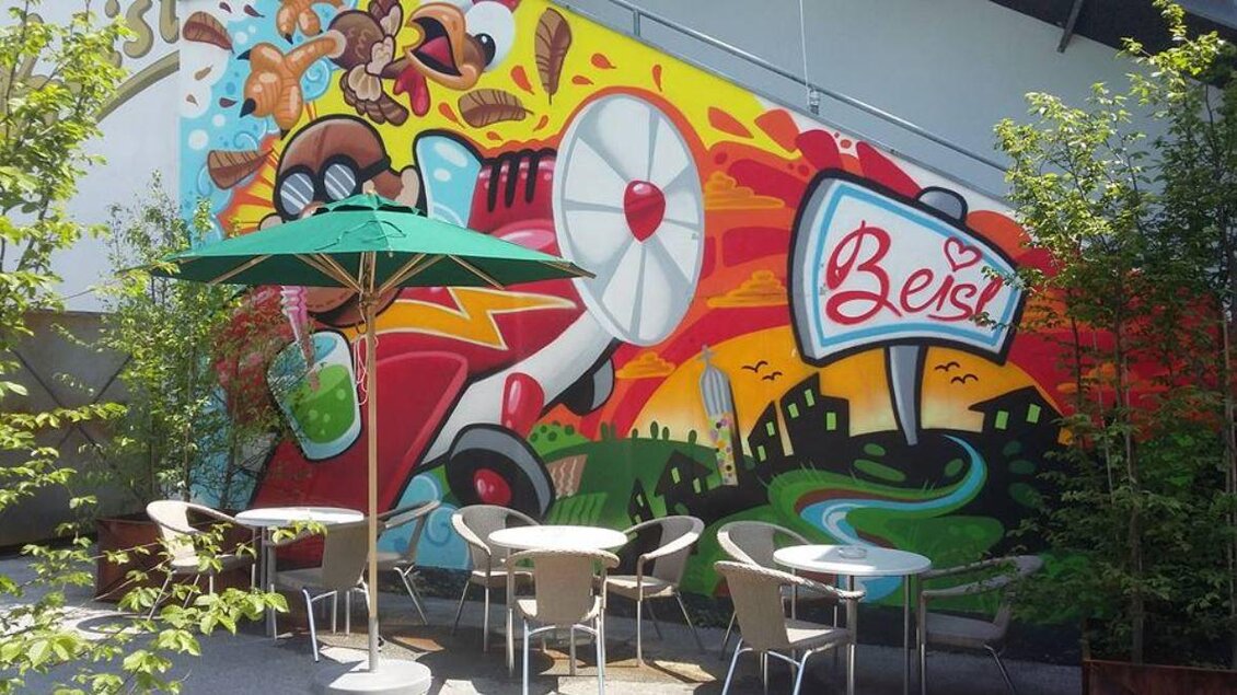 Ein buntes Wandgemälde mit einem Flugzeug und einer schönen Landschaft. Im Vordergrund stehen Tische und Stühle unter einem Sonnenschirm. | © Café Beisl Feldbach