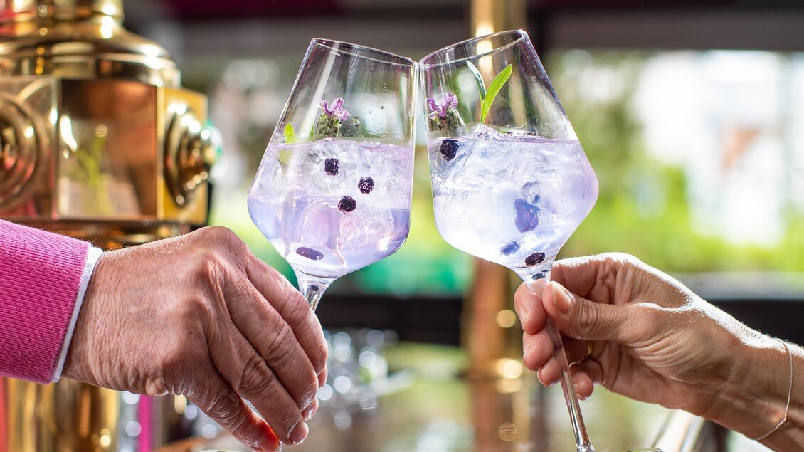 Zwei Gläser mit lila Getränken werden zum Anstoßen erhoben. Im Hintergrund ist eine elegante Bar mit goldenen Akzenten zu sehen. | © Cafe Beisl