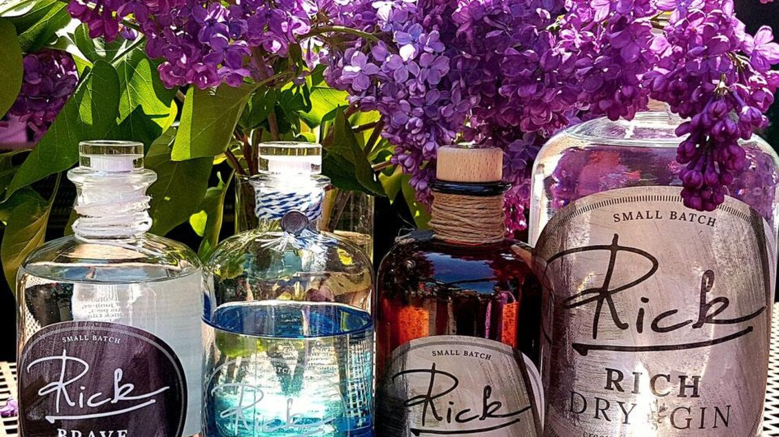 Vier Flaschen Gin mit dem Namen "Rick" stehen vor blühenden lila Fliederblüten. Die Flaschen haben unterschiedliche Designs und Farben. | © Café Beisl Feldbach