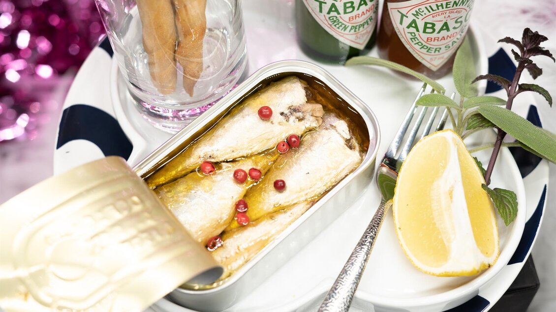 Ein Teller mit einer offenen Sardinendose, die mit rosa Pfefferkörnern gefüllt ist. Daneben befinden sich ein Glas, eine Zitronenscheibe und zwei Flaschen Tabasco. | © Cafe Beisl