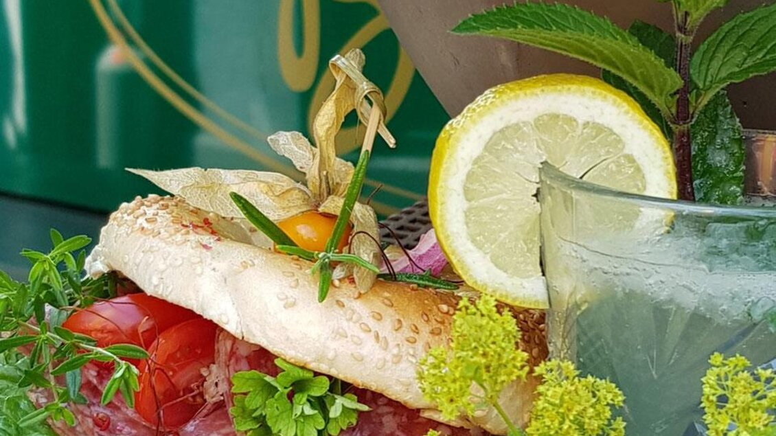 Ein frisch zubereitetes Sandwich mit Salami, Tomaten und einer grünen Salatbasis. Daneben steht ein Getränk mit einer Zitronenscheibe und frischen Kräutern. | © Café Beisl Feldbach