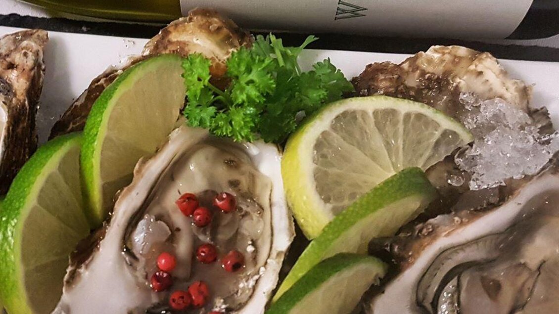 Frische Austern auf einem Teller, garniert mit Limettenscheiben und Pfefferkörnern. Im Hintergrund steht eine Flasche Weißwein. | © Café Beisl Feldbach