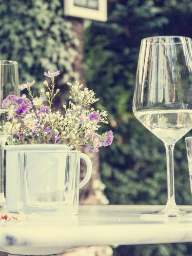 A beautiful outdoor table with glasses and fresh flowers. In the background, a sign with information is visible. | © Cafe Fotter