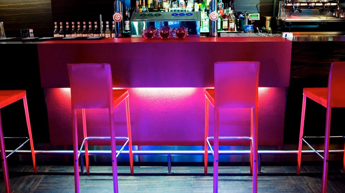 Eine moderne Bar mit leuchtend roten Barhockern und einer stilvollen Theke. Im Hintergrund sind verschiedene Getränke und Gläser sichtbar. | © Cafe Glockenspiel