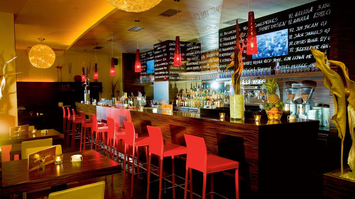 Eine stilvolle Bar mit roten Stühlen und einer langen Bartheke. Die Wände sind mit Getränkekarten dekoriert und es gibt stimmungsvolle Beleuchtung. | © Cafe Glockenspiel