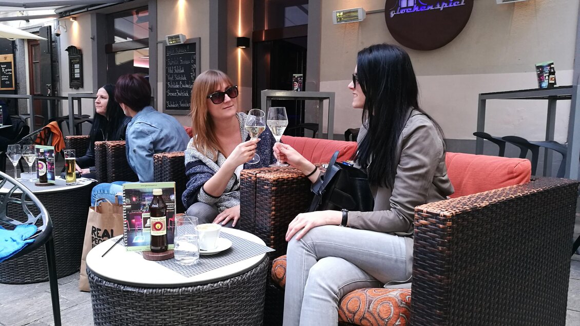Zwei Frauen sitzen entspannt in einem Café und stoßen mit ihren Gläsern an. Um sie herum sind weitere Gäste in einem gemütlichen Ambiente. | © Cafe Glockenspiel