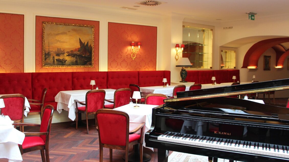 Eleganter Raum mit roten Samtsesseln, gedeckten Tischen, glitzerndem Kronleuchterlicht und schwarzem Flügel, der das Ambiente prägt | © Region Graz - Vanessa Züttl