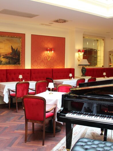 Elegant room with red velvet chairs, set tables, sparkling chandelier light, and a black grand piano that shapes the ambiance | © Region Graz - Vanessa Züttl