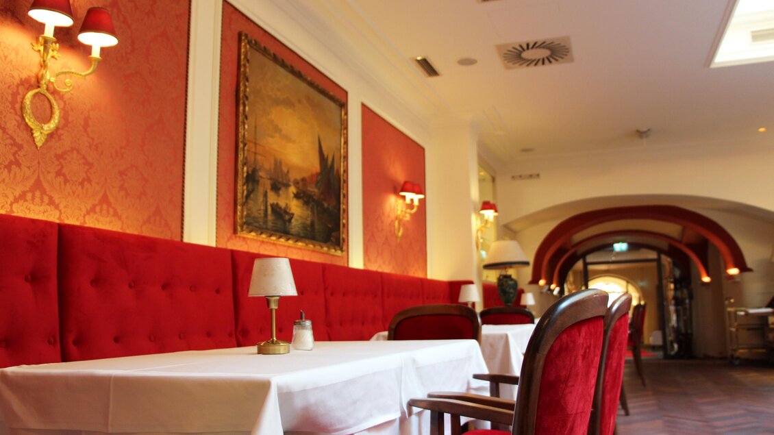 Gastraum mit roten Samtpolstern, weißen Tischdecken, Wandlampen in Gold und klassischem Gemälde vor Arkaden im Hintergrund | © Region Graz - Vanessa Züttl