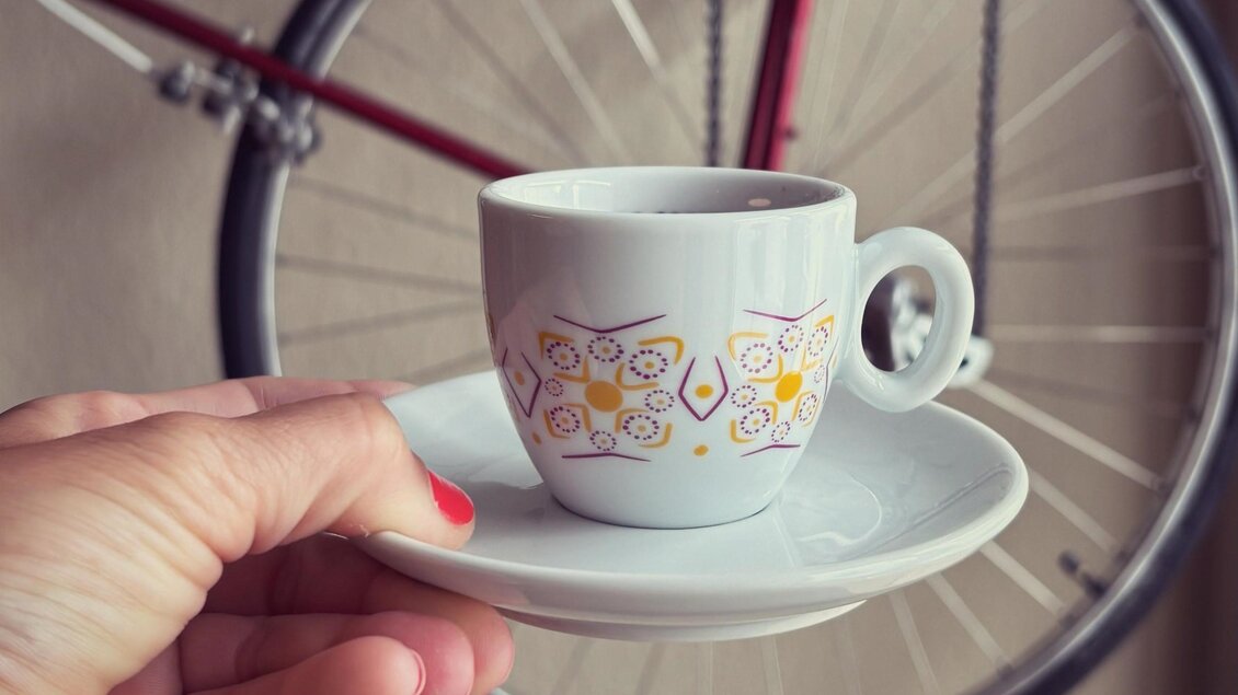 Eine Hand hält eine elegante Tasse mit Untertasse. Im Hintergrund ist ein Fahrradrad zu sehen. | © Café Hirzenberger