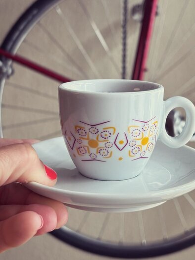 Eine Hand hält eine elegante Tasse mit Untertasse. Im Hintergrund ist ein Fahrradrad zu sehen. | © Café Hirzenberger