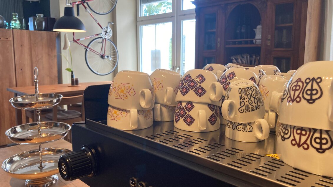 Eine Übersicht von Kaffeetassen auf einer Espresso-Maschine. Im Hintergrund sind Fahrräder und eine gemütliche Einrichtung zu sehen. | © Café Hirzenberger