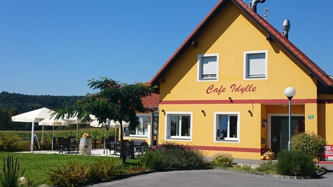 Ein gelbes Café mit dem Namen "Café Idylle" in ländlicher Umgebung. Vor dem Gebäude gibt es Sitzgelegenheiten im Freien und eine grüne Landschaft im Hintergrund. | © Café Idylle