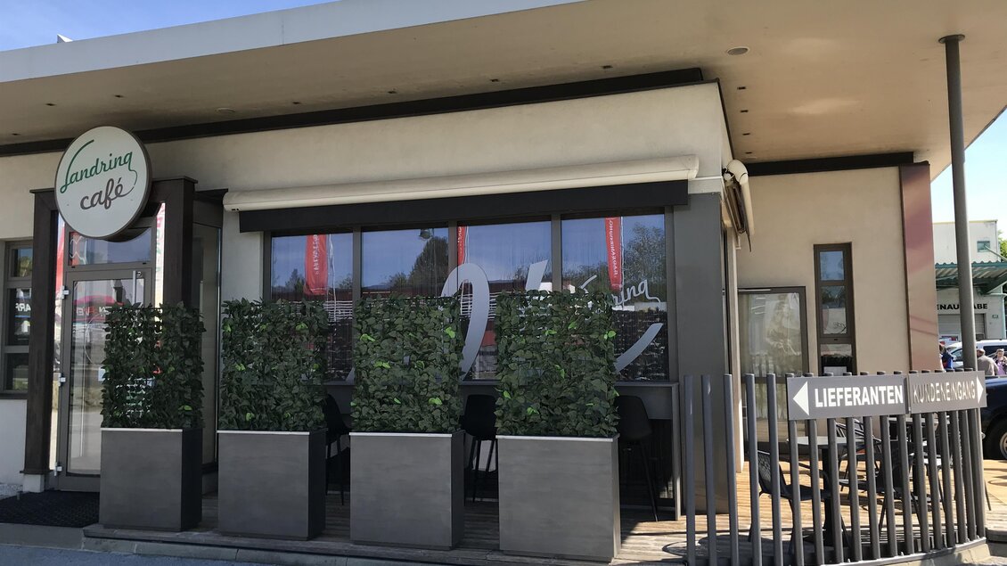 Ein modernes Café mit großen Fenstern und einem einladenden Eingang. Grünpflanzen umgeben die Terrasse und ein Schild weist auf Lieferdienste hin. | © Tourismusverband Oststeiermark