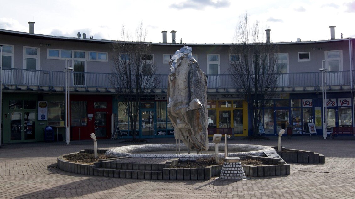 Ein moderner Brunnen mit einer stilisierten Statue in der Mitte. Im Hintergrund sind verschiedene Geschäfte in einem offenen Bereich zu sehen. | © Gemeinde Gratkorn - Gasser