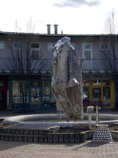 Ein moderner Brunnen mit einer stilisierten Statue in der Mitte. Im Hintergrund sind verschiedene Geschäfte in einem offenen Bereich zu sehen. | © Gemeinde Gratkorn - Gasser
