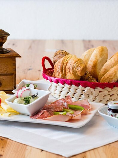 Ein Frühstückstisch mit frischen Brötchen, Wurst, Käse und einer Obstschale. Dazu gibt es frisch gepressten Saft und eine Kaffeemühle als Dekoration. | © Facebook