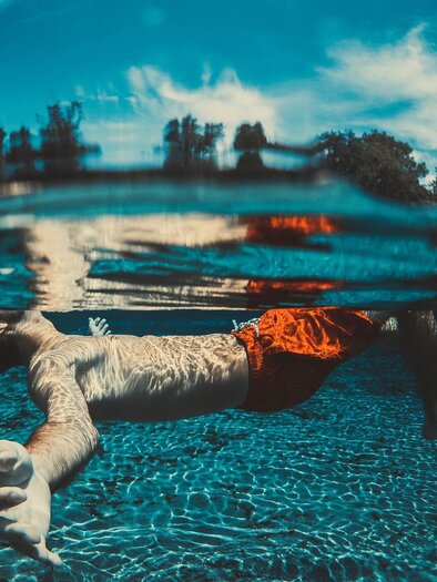 Ein Schwimmer liegt entspannt im Wasser. Die Oberfläche reflektiert das Licht und umgibt ihn mit einem klaren, blauen Himmel im Hintergrund. | © Symbolbild PixaBay / Pexels