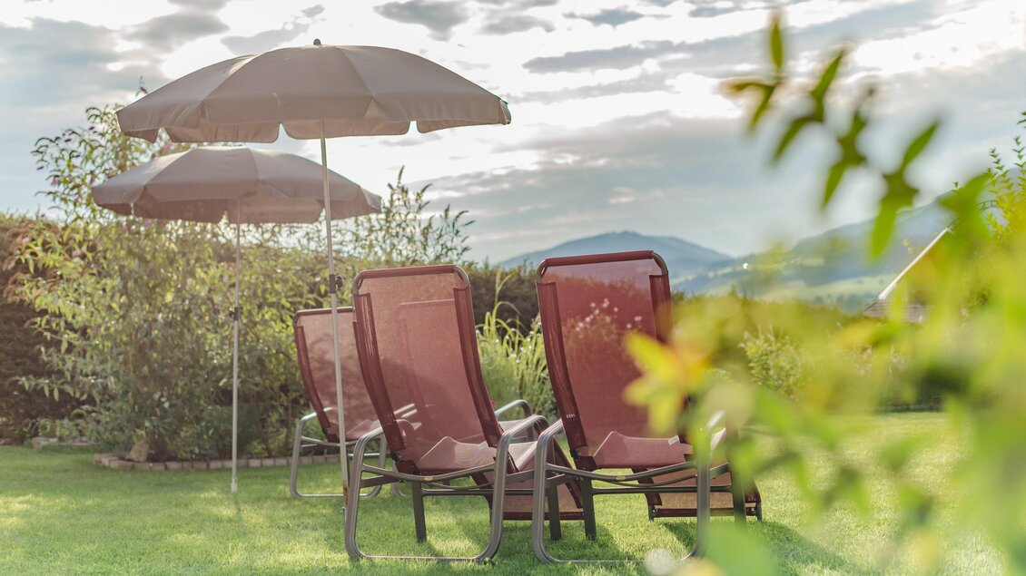 Eine entspannte Gartenszene mit zwei Liegestühlen und einem Sonnenschirm. Im Hintergrund sind sanfte Hügel und ein bewölkter Himmel zu sehen. | © Christine Höflehner