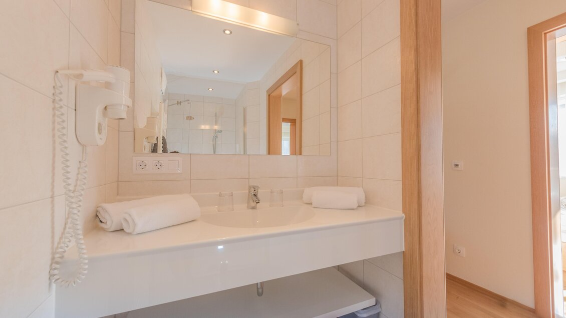 Ein modernes Badezimmer mit einem weißen Waschbecken und einem großen Spiegel. Frische Handtücher liegen bereit und das Licht ist freundlich. | © Christine Höflehner