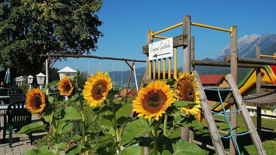 Ein Spielplatz mit einer Schaukel und bunten Rutschen. Im Vordergrund blühen sonnige Sonnenblumen unter einem klaren blauen Himmel. | © Café Pension Kitzer