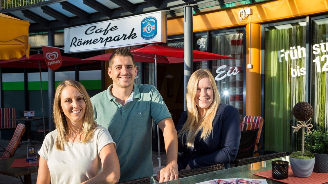 Drei freundliche Mitarbeiter stehen vor dem Café Römerpark. Im Hintergrund sind Sonnenschirme und das Café-Schild zu sehen. | © TV Region Graz - René Vidalli