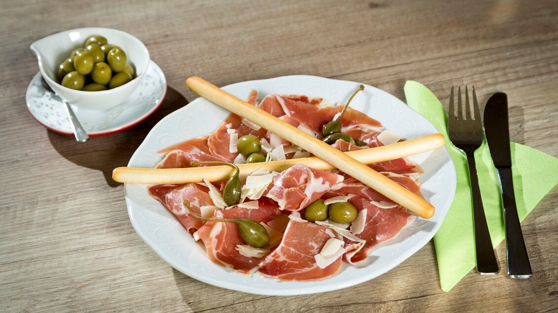 Ein Teller mit geschnittenem Schinken, garniert mit grünen Oliven und Grissini. Daneben eine Schale mit zusätzlichen grünen Oliven. | © TV Region Graz - René Vidalli