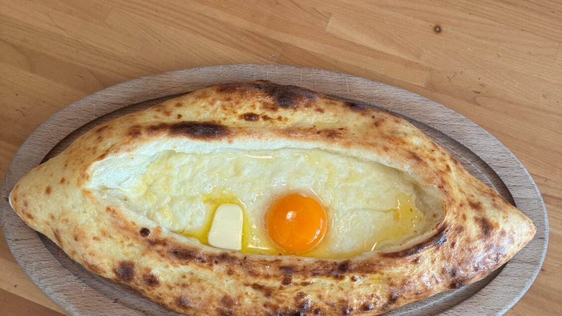 Ein frisch gebackenes Khachapuri auf einem Holzbrett. Es ist gefüllt mit Käse, einem Stück Butter und einem rohen Ei in der Mitte. | © Café Restaurant Freischwimmer