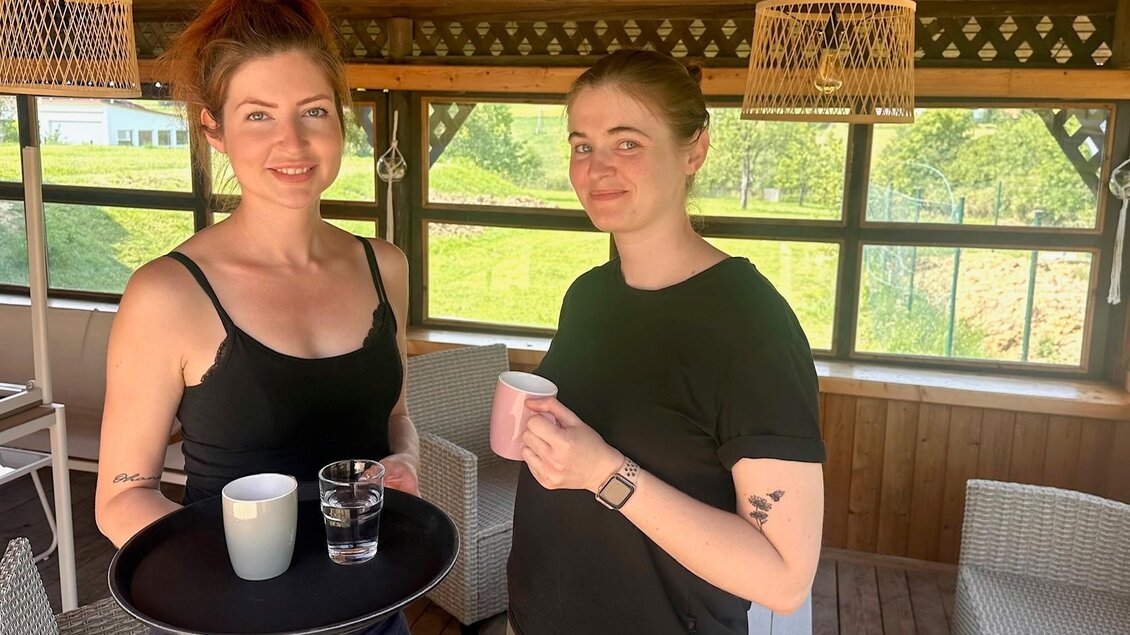 Zwei Frauen stehen in einem gemütlichen Raum und halten ein Tablett mit Getränken. Der Raum hat große Fenster und ist mit hellen Möbeln ausgestattet. | © Café Sophia