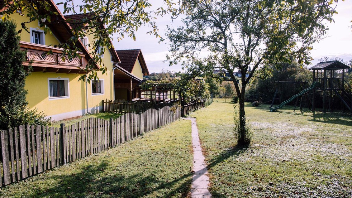 Ein gelbes Haus mit einem gepflegten Garten und einem Weg. Im Hintergrund ist ein Spielplatz mit Spielgeräten und ein großer Baum zu sehen. | © Kurkommission Bad Blumau