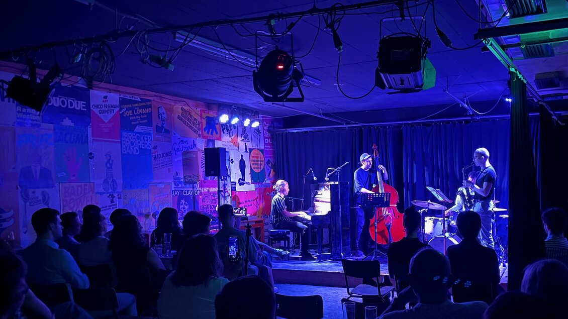 Eine jazzige Live-Performance in einem kleinen Club mit einer Band aus Pianist, Bassist und Schlagzeuger. Das Publikum genießt die Musik in einer stimmungsvollen, beleuchteten Umgebung. | © Cafe Stockwerk - N.Popp