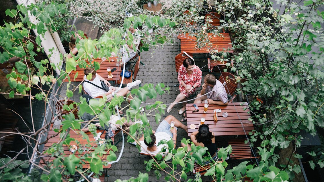Eine gemütliche Außenterrasse mit Holztischen, umgeben von Pflanzen. Fünf Personen sitzen zusammen und genießen ihre Zeit. | © Cafe Stockwerk - G.Obetzhofer
