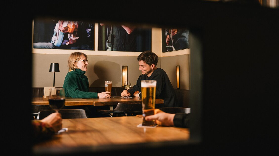 Zwei Personen sitzen an einem Tisch in einem gemütlichen Lokal und unterhalten sich. Im Hintergrund sind Fotos an der Wand sowie Biergläser zu sehen. | © Cafe Stockwerk - G.Obetzhofer