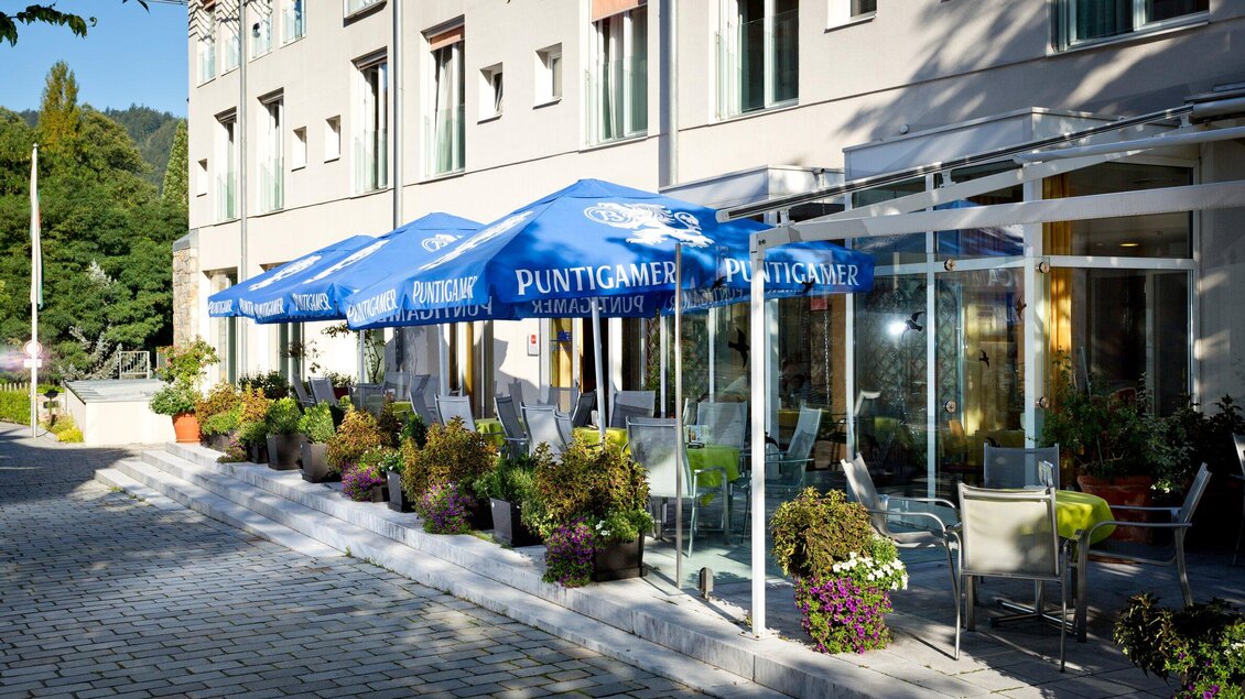 Ein modernes Café mit blauen Schirmen und einer einladenden Terrasse. Die Umgebung ist grün und gepflegt, ideal für entspannte Stunden im Freien. | © TV Region Graz - René Vidalli