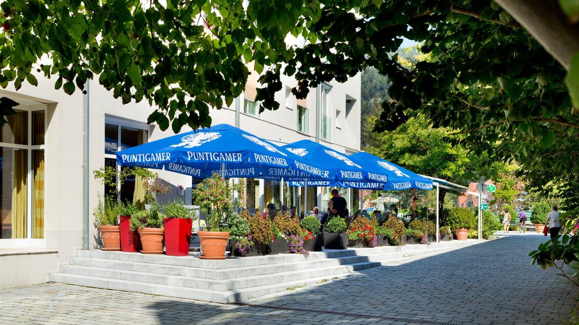 Ein gemütliches Café mit blauen Sonnenschirmen und bunten Pflanzen. Die Umgebung ist einladend und gut beleuchtet. | © TV Region Graz - René Vidalli