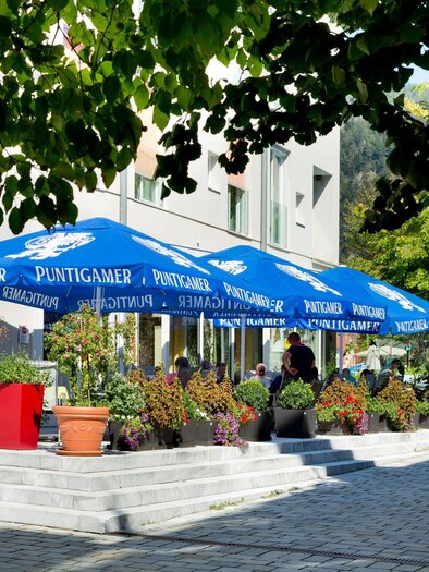Ein gemütliches Café mit blauen Sonnenschirmen und bunten Pflanzen. Die Umgebung ist einladend und gut beleuchtet. | © TV Region Graz - René Vidalli