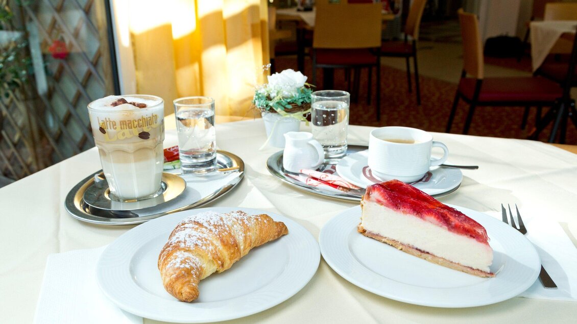 Ein gemütliches Frühstück mit einem Croissant, einem Stück Käsekuchen mit Fruchtbelag, einer Tasse Kaffee und einem Glas Wasser. Der Tisch ist hell dekoriert und lädt zum Verweilen ein. | © TV Region Graz - René Vidalli