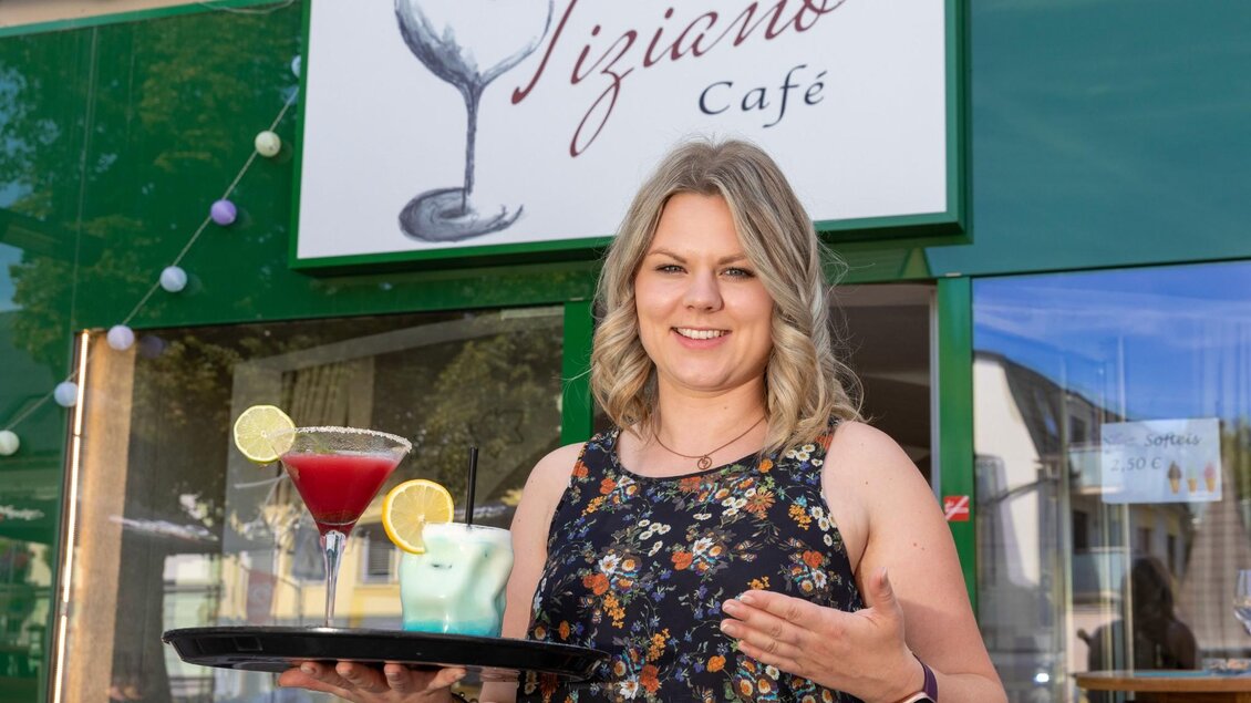 Eine freundliche Kellnerin steht vor dem Tiziano Café und hält ein Tablett mit Cocktails. Im Hintergrund ist das Café-Schild gut sichtbar. | © TV Region Graz - Harry Schiffer