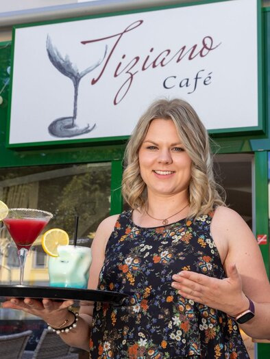 Eine freundliche Kellnerin steht vor dem Tiziano Café und hält ein Tablett mit Cocktails. Im Hintergrund ist das Café-Schild gut sichtbar. | © TV Region Graz - Harry Schiffer