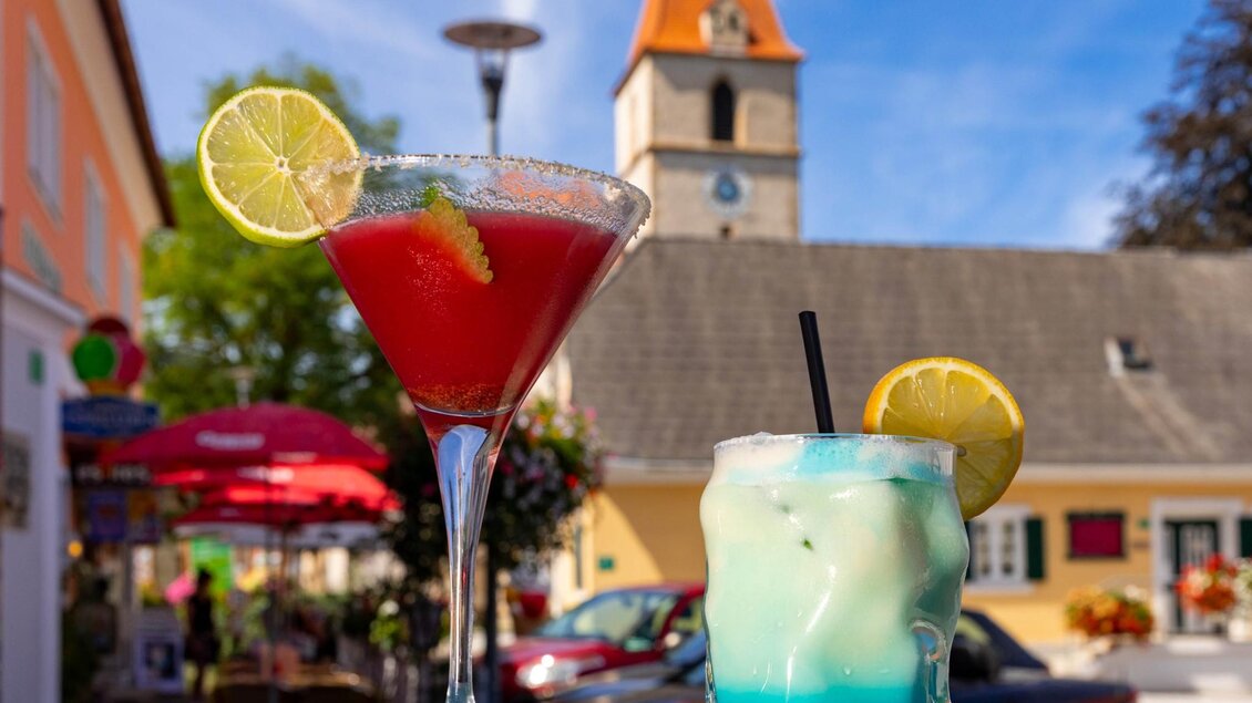 Zwei Cocktails stehen auf einem Tisch, einer rot und einer blau-weiß. Im Hintergrund ist ein Kirchturm und eine lebhafte Straßenszene mit Tischen und Sonnenschirmen zu sehen. | © TV Region Graz - Harry Schiffer