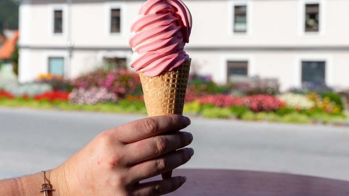 Hand hält ein Softeis im Café Tiziano in Semriach, im Hintergrund blühende Beete und Hausfassade | © TV Region Graz - Harry Schiffer