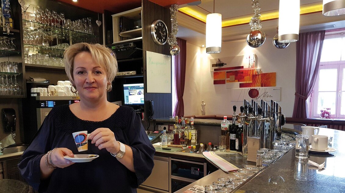 Eine Frau steht an einer Bar und hält eine Tasse Kaffee in der Hand. Im Hintergrund sind Glasregale und eine Bar mit Getränken zu sehen. | © Josy Lammers