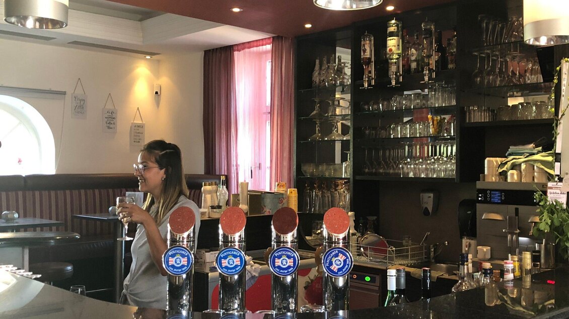 Eine Frau steht an der Bar und hält ein Getränk in der Hand. Im Hintergrund sind Gläser und Flaschen auf einem Regal sichtbar. | © Josy Lammers