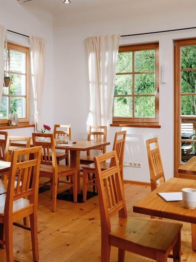 A bright and inviting café with wooden tables and chairs. Large windows provide a view of greenery. | © Viola im Garten