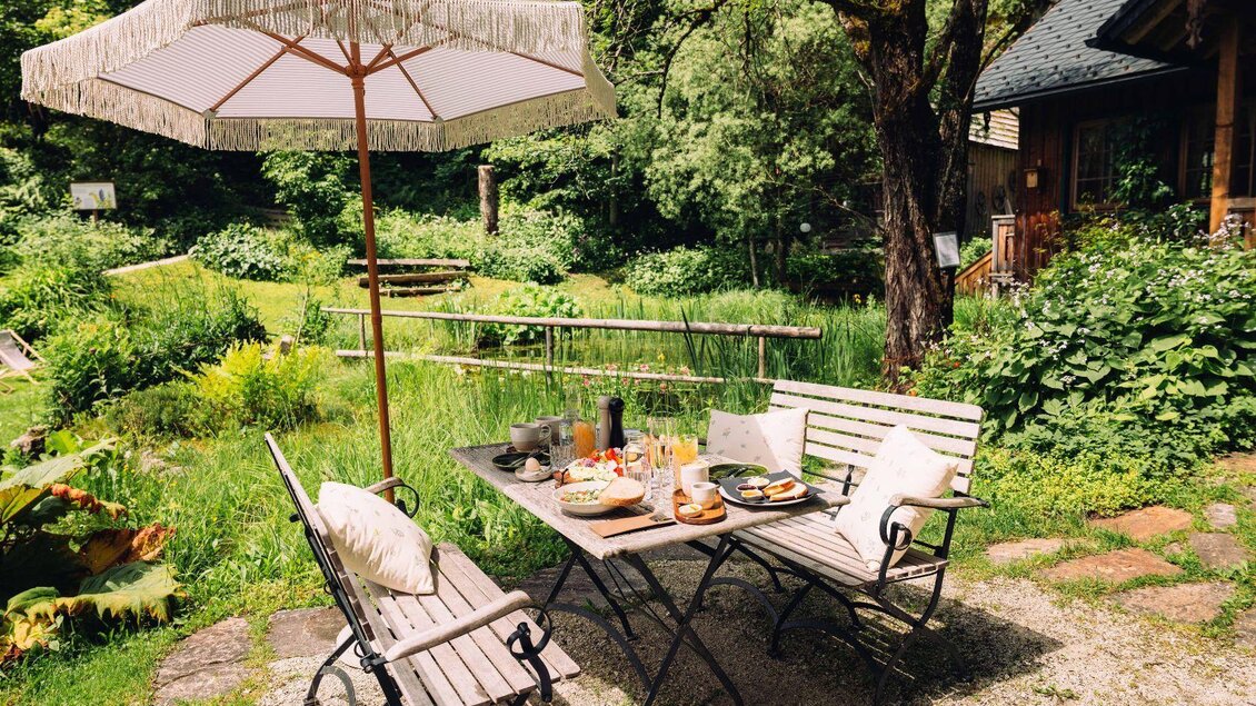 Ein schöner Außenbereich mit einem Tisch und zwei Stühlen unter einem Sonnenschirm. Der Tisch ist mit Essen dekoriert, umgeben von einer grünen Gartenlandschaft. | © Viola im Garten