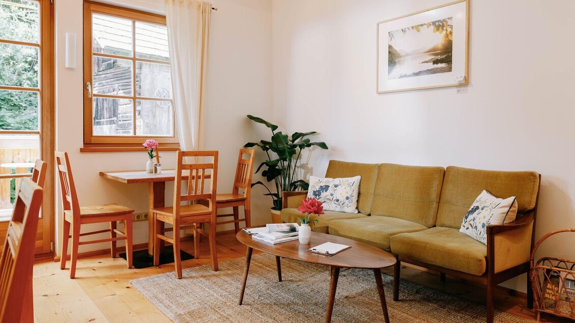 Ein gemütliches Wohnzimmer mit einem grünen Sofa und einem Holztisch. Große Fenster lassen viel Licht herein und eine Pflanze sorgt für einen frischen Akzent. | © Viola im Garten