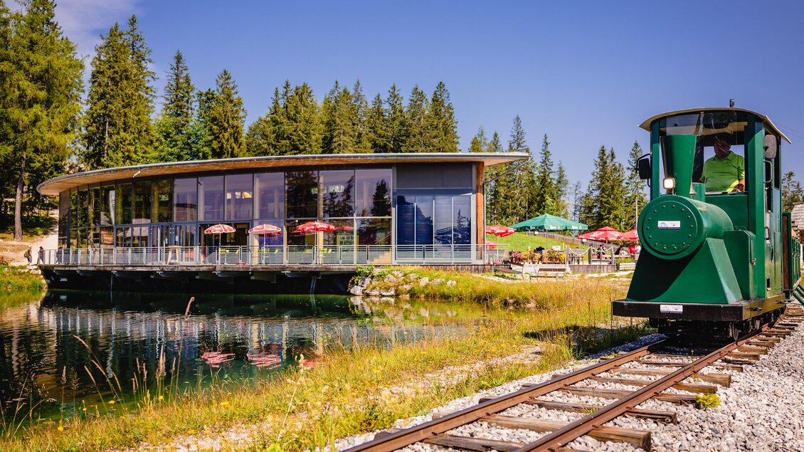 Eine grüne Eisenbahn an einem Teich, neben einem modernen Gebäude mit großen Fenstern. Im Hintergrund sind Bäume und einige Tische mit Sonnenschirmen zu sehen. | © Mariazeller Bürgeralpe / Rudy Dellinger
